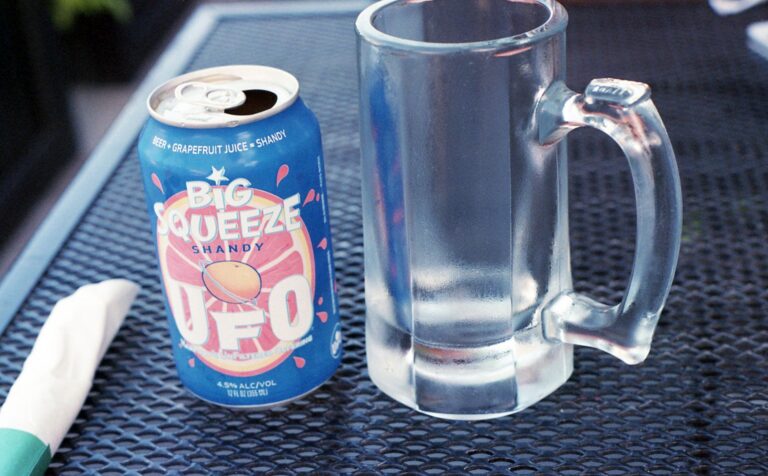 A vibrant shandy beer can and frosted mug on a metal table outdoors.