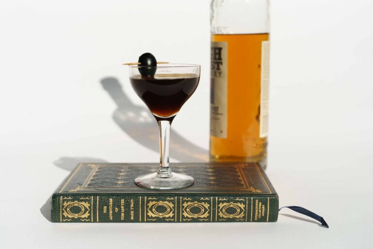 Composition of glass bottle of whiskey near Manhattan cocktail served with pickled olive on stick on book